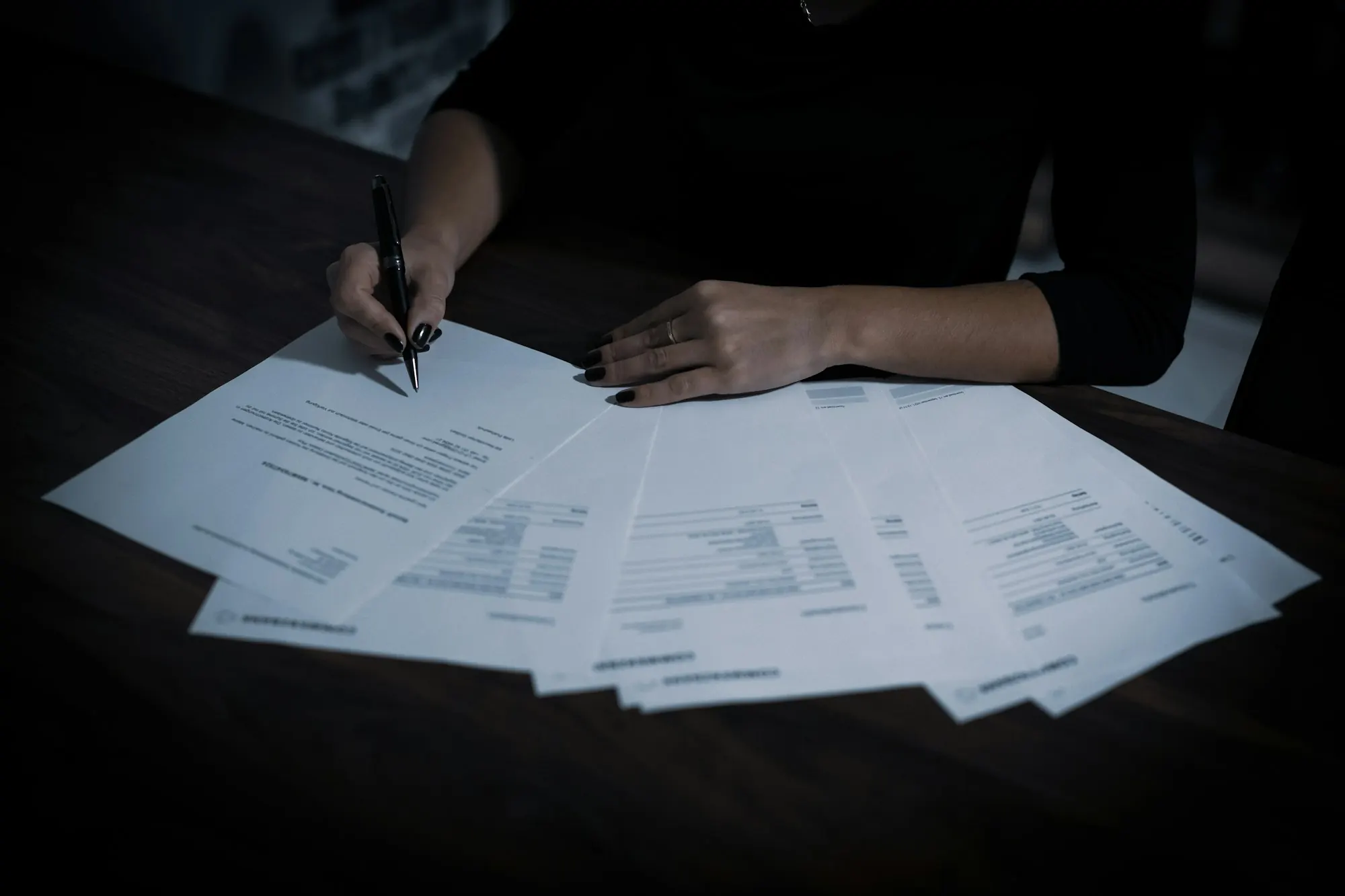 Hands signing paperwork spread across a dark table
